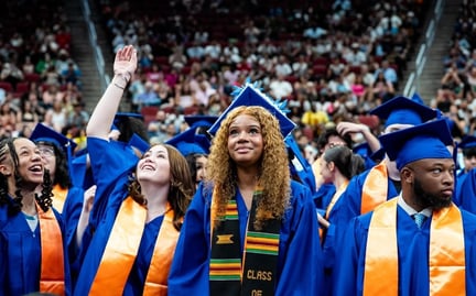 Students in blue graduation caps and gowns are standing together, with one student raising her hand and others smiling