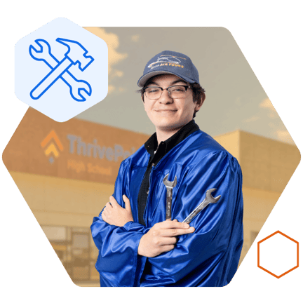 A student is wearing a graduation gown and cap while holding two wrenches and smiling in front of a school building