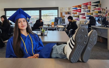 A student in a blue graduation cap and gown is sitting at a desk with her feet up and looking at the camera in a classroom